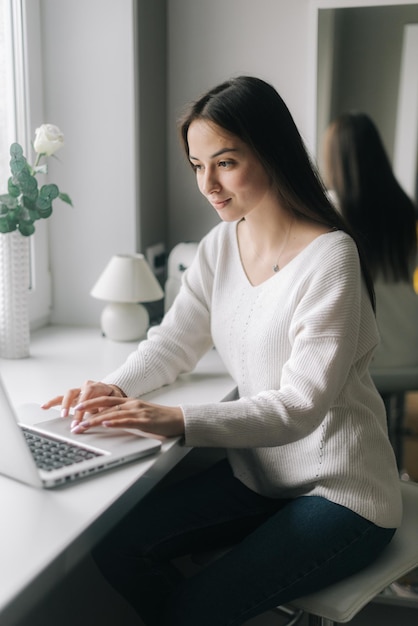 Una giovane donna freelance seria e concentrata sta digitando sul laptop seduto accanto a una finestra in cucina Concetto di vita quotidiana