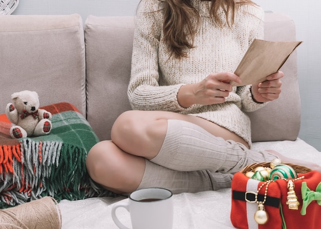 Una giovane donna graziosa con un maglione leggero si siede su un letto con decorazioni natalizie e legge una lettera