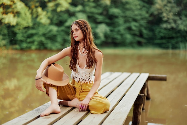 Una giovane donna hippie si siede su un ponte sul lago indossando eleganti abiti ecologici e sorridendo