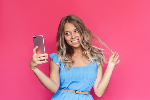 Una giovane donna in abito blu si fa un selfie con il suo cellulare su uno sfondo rosa brillante