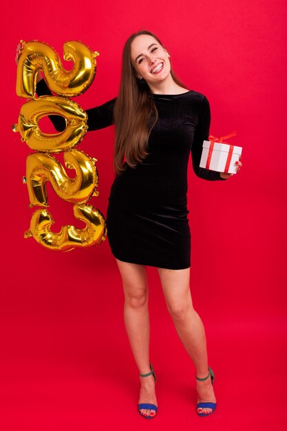 Una giovane donna in abito nero e tacchi con una confezione regalo tiene le figure del nuovo anno 2023 su sfondo rosso