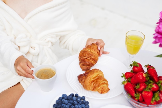 Una giovane donna in accappatoio e un asciugamano in testa si gode la vacanza al resort e fa colazione caffè con croissant e fragole