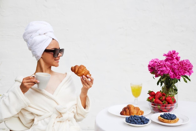 Una giovane donna in accappatoio e un asciugamano in testa si gode la vacanza al resort e fa colazione con caffè con croissant e fragole