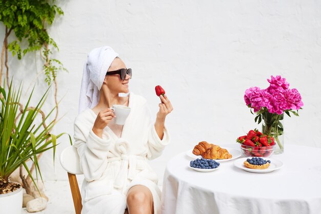 Una giovane donna in accappatoio e un asciugamano sulla testa mangia fragole si gode la sua vacanza al resort fa colazione caffè con un croissant e fragole