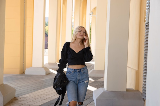 Una giovane donna in camicia e jeans cammina per strada e parla al telefono