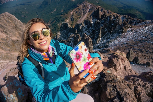 Una giovane donna in giacca blu che fa foto con il suo smartphone nella zona di montagna durante la stagione fredda con nuvole sullo sfondo