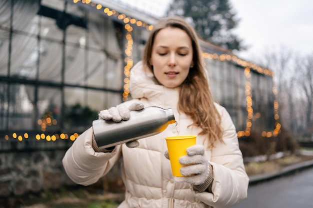 Una giovane donna in giacca invernale versa il tè da un thermos in un bicchiere di carta mentre si trova sullo sfondo di un edificio con una ghirlanda Trascorrere del tempo all'aperto in inverno