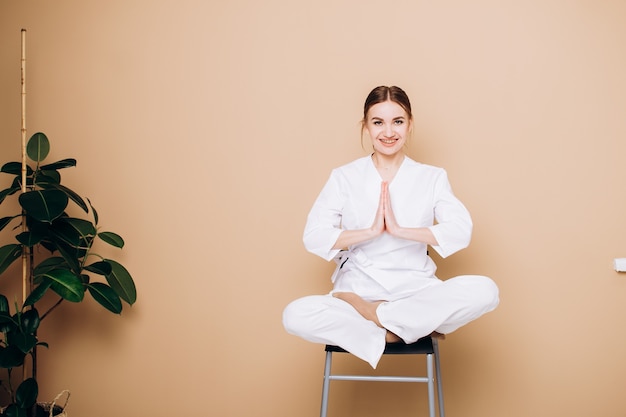 Una giovane donna in kimono bianco si siede su una sedia e medita in una posa yoga a casa