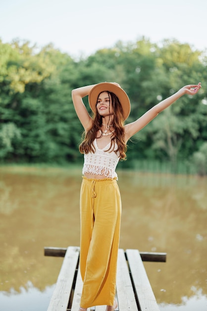 Una giovane donna in look hippie e ecodress viaggia nella natura in riva al lago indossando un cappello e pantaloni gialli al tramonto autunnale