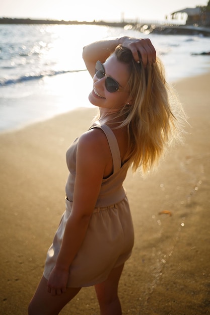 Una giovane donna in occhiali da sole in un top beige e pantaloncini corti cammina lungo la riva del mare al tramonto