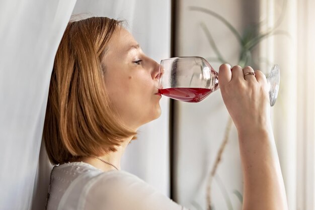 Una giovane donna in piedi alla finestra beve da un bicchiere di vino rosso