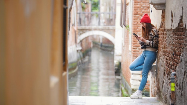 Una giovane donna in piedi vicino al canale dell'acqua e alla ricerca di qualcosa nel suo telefono Venezia Italia