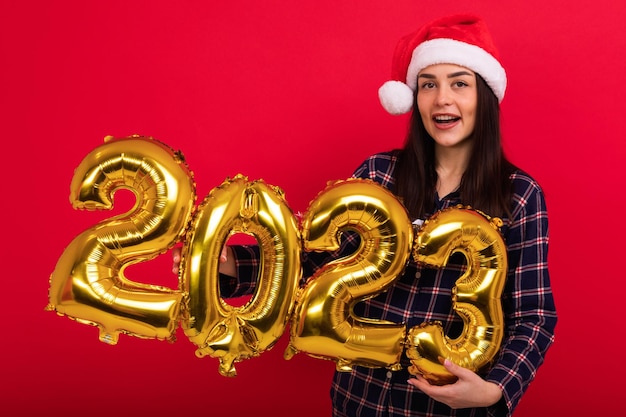 Una giovane donna in pigiama e un cappello di Babbo Natale tiene palloncini a forma del nuovo anno 2023 Sfondo rosso studio