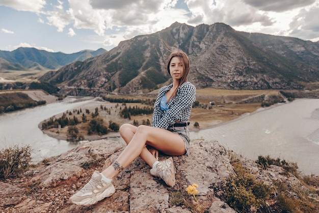 Una giovane donna in posa seduta a gambe incrociate sulle rocce grigie sullo sfondo di montagne rocciose e un chiaro ruscello che scorre in lontananza. Vista pittoresca. Sessione fotografica sulla natura aperta.