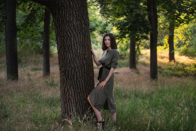 Una giovane donna in un abito verde kaki sta con la schiena contro un albero al tramonto nel parco.