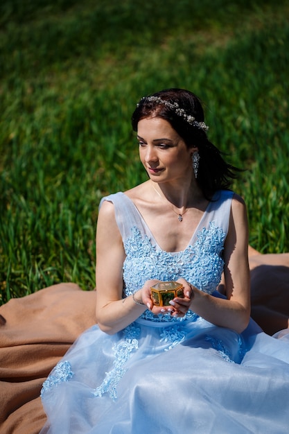 Una giovane donna in un lungo abito blu siede contro un campo verde con una bara tra le mani. Ritratto di moda di una bella ragazza con un sorriso sul viso. Scatola dell'anello di fidanzamento