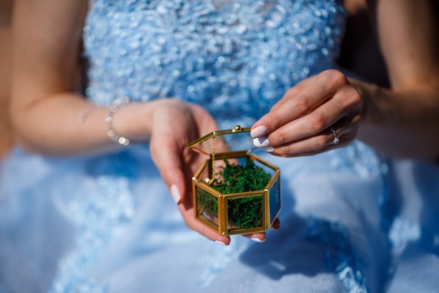 Una giovane donna in un lungo abito blu siede contro un campo verde con una bara tra le mani. Ritratto di moda di una bella ragazza con un sorriso sul viso. Scatola dell'anello di fidanzamento