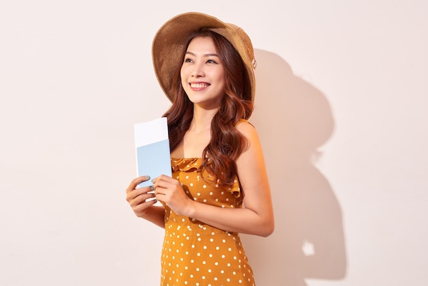 Una giovane donna in un vestito arancione a pois con un cappello di paglia in testa ha in mano un passaporto e dei biglietti aerei