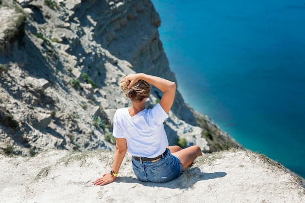 Una giovane donna in una maglietta bianca siede su un'alta scogliera sul mare Natura magnifica Stile di vita attivo energia e salute