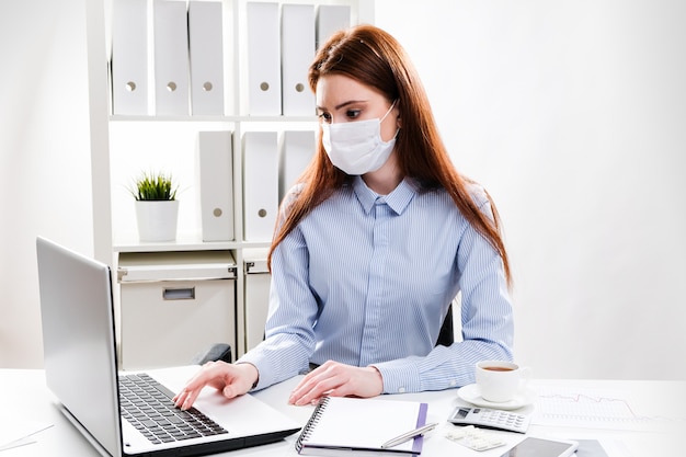 Una giovane donna in una maschera protettiva lavora su un computer. Donna di affari in una mascherina medica in ufficio.