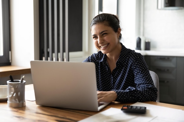Una giovane donna indiana sorridente si siede alla scrivania a casa, gestisce le spese domestiche, paga online sul portatile, una donna etnica millenaria felice fa pagamenti su Internet sul computer, un concetto di risparmio.