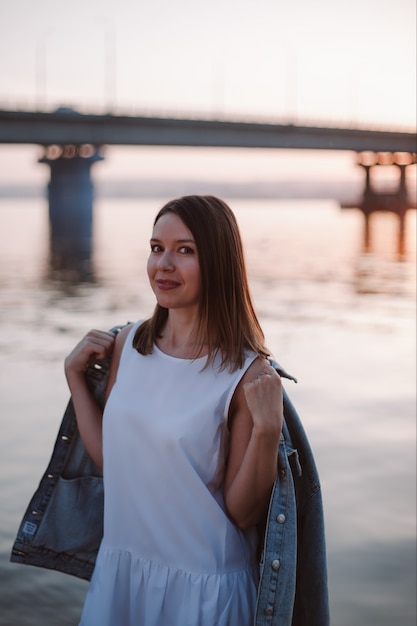 Una giovane donna indossa una giacca di jeans in una fresca serata estiva al tramonto vicino al ponte sul fiume