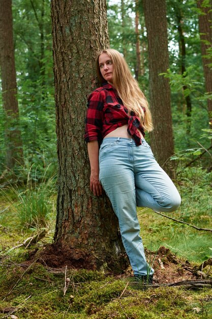 Una giovane donna italiana che indossa una flanella rossa con dei jeans e si appoggia a un albero in una foresta