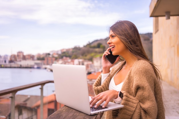 Una giovane donna latina con una maglietta bianca e un maglione di lana marrone che lavora in telelavoro sul terrazzo di una casa in riva al mare parlando al telefono