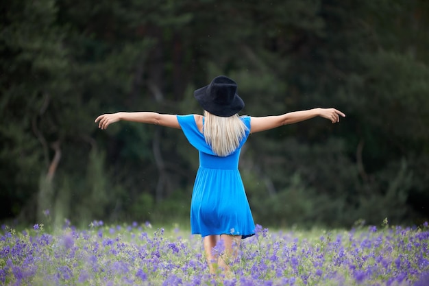 Una giovane donna magra in un vestito blu con un cappello nero cammina in un campo di fiori in estate