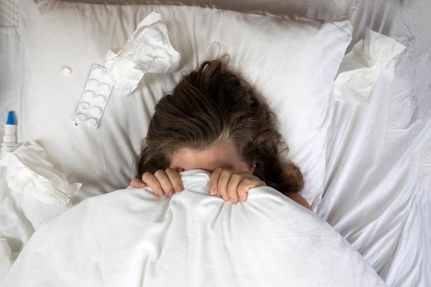 Una giovane donna malata a letto, sdraiata con la testa sotto le coperte e un mucchio di fazzoletti accanto a lei