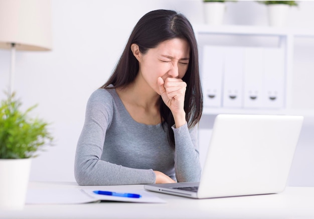 Una giovane donna malata impegnata al computer portatile lotta contro la tosse con il coronavirus o l'influenza sul posto di lavoro