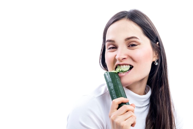 Una giovane donna mangia un cetriolo Bella bruna sorridente in un maglione bianco Cibo sano e vegetarismo Isolato su sfondo bianco Spazio per il testo