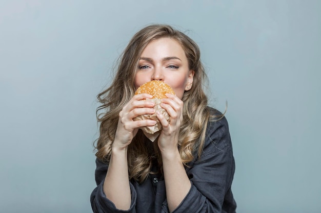 Una giovane donna mangia un hamburger con piacere Bella bionda sorridente in pigiama contro un muro grigio Cibo malsano a letto
