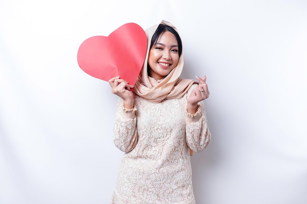 Una giovane donna musulmana asiatica felice che indossa un hijab sente il gesto del cuore di forme romantiche che esprime sentimenti teneri e tiene in mano una carta rossa a forma di cuore