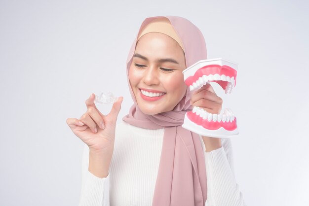 Una giovane donna musulmana che tiene bretelle invisalign e un modello dentale artificiale di uomo su bianco, assistenza sanitaria dentale e concetto ortodontico.