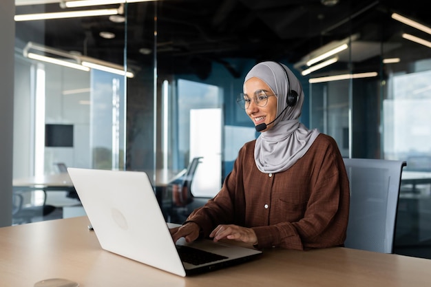 Una giovane donna musulmana in hijab e cuffie sta lavorando in ufficio a una scrivania usando un portatile