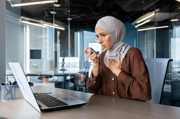 Una giovane donna musulmana in hijab è seduta a una scrivania in ufficio e usando un inalatore ha un