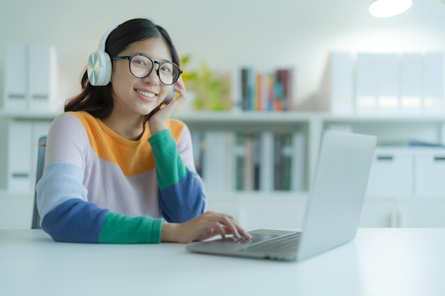 Una giovane donna o uno studente che utilizza un computer portatile in biblioteca mentre indossa occhiali e cuffie Sorride e sembra felice