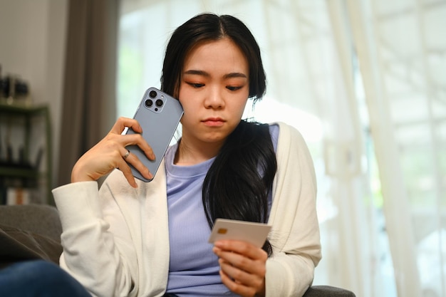 Una giovane donna preoccupata che tiene in mano smartphone e carta di credito guarda seriamente e si preoccupa per l'angoscia