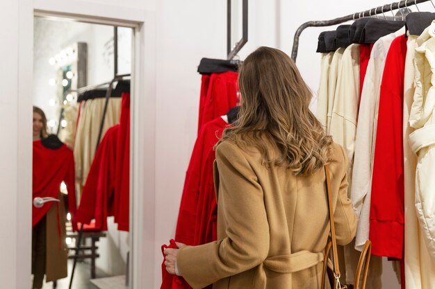 Una giovane donna prova un vestito davanti a uno specchio in un negozio di abbigliamento. Bella bionda sorridente in un cappotto beige.