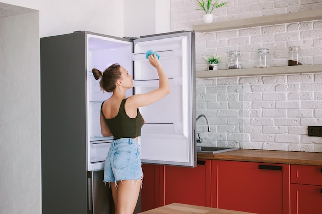 Una giovane donna pulisce il frigorifero con uno straccio in cucina Faccende intimità e comfort