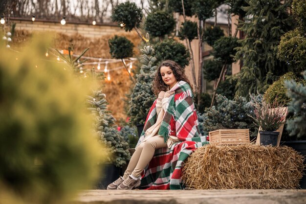 Una giovane donna riccia avvolta in un caldo plaid in una cella siede in attesa del nuovo anno La ragazza sullo sfondo del verde degli alberi di Natale sulla strada Atmosfera festiva