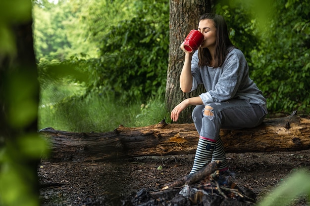 Una giovane donna si scalda vicino a un fuoco spento con una tazza di bevanda calda nella foresta tra gli alberi
