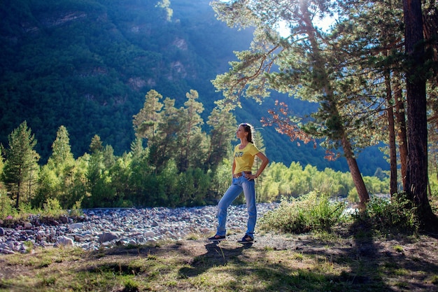 Una giovane donna si trova sulla riva di un fiume di montagna ai margini di una foresta e ammira la natura.