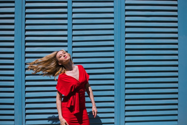 Una giovane donna si trova vicino a un muro con persiane blu Luce dura di una giornata di sole brillante La ragazza sviluppa i capelli