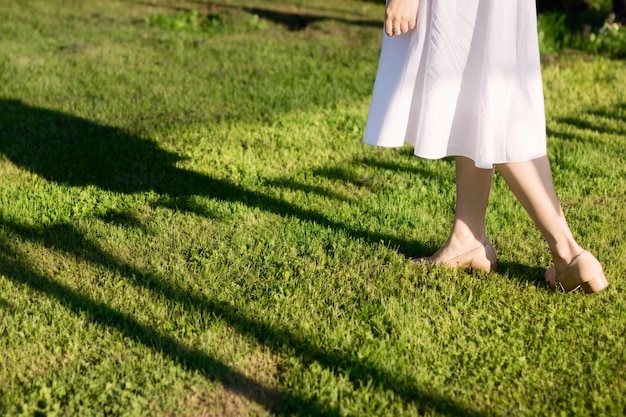 Una giovane donna snella con un vestito bianco e scarpe beige cammina sull'erba verde brillante Solo le sue gambe sono nel framexA