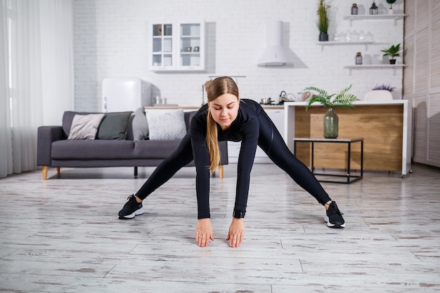 Una giovane donna snella fa sport a casa, indossando abiti da fitness, un top nero e leggings. Fitness a casa per un bel corpo. Riscaldati prima di fare sport