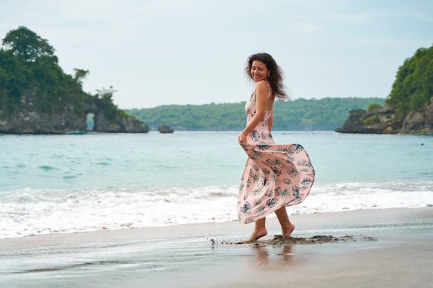 Una giovane donna snella in un vestito rosa chiaro cammina lungo una spiaggia tropicale la ragazza sta ballando nelle onde del mare