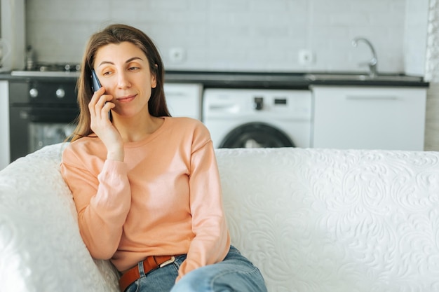 Una giovane donna soddisfatta che si gode un pasto nella sua cucina accogliente mentre parla con sua sorella sul suo smartphone