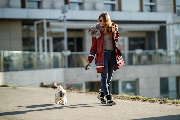 Una giovane donna sorridente che cammina con il suo simpatico cane Shih Tzu per la strada della città.